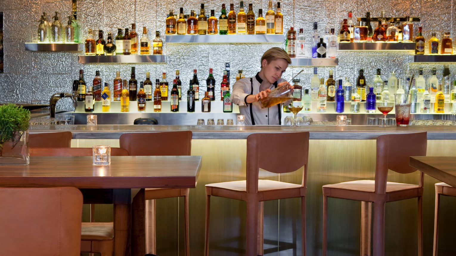 Pony Bar server pours cocktail into glass behind wood bar with stools, in front of wall of liquor bottles