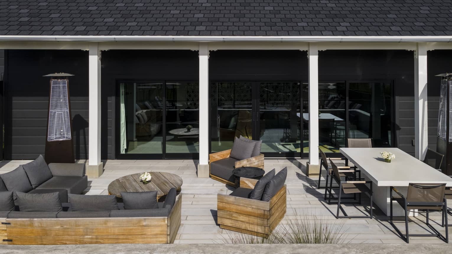 Luxury villa patio with gray cushioned sofa, wooden armchairs, a round coffee table, rectangular dining table and outdoor heaters against a black exterior wall