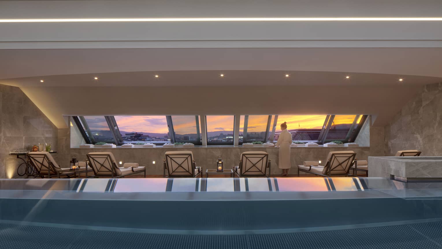 Woman in robe stands on deck beside blue indoor swimming pool, lounge chairs, looking at sunset through window