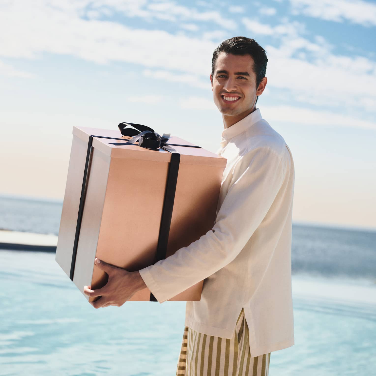 Man wearing white shirt and striped pants carries large present, with the ocean in the background