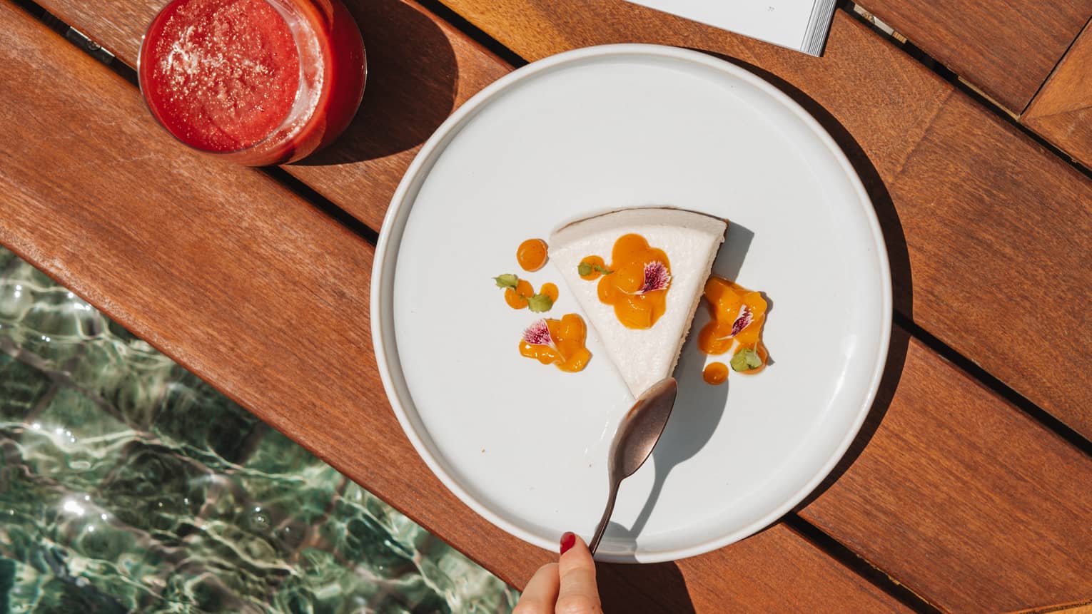A wood surface with an open book placed on it next to a reddish drink in a tumbler and a white plate holding a white and orange food item. A person reaches for a bite of the food with a spoon.