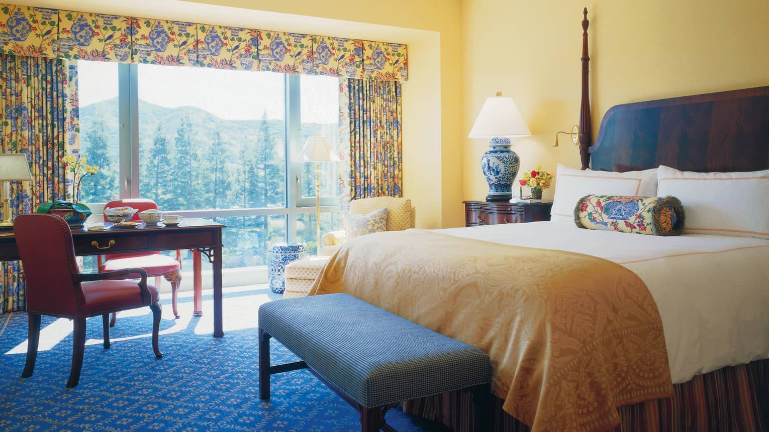 Deluxe Room hotel bed with gold blanket, bench, desk, windows with bright curtains