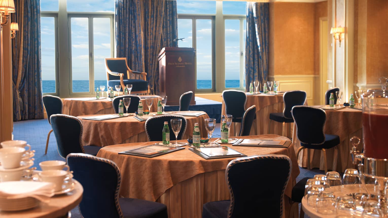 Sun-lit meeting room with round tables, royal blue chairs, podium facing floor-to-ceiling window and ocean views 