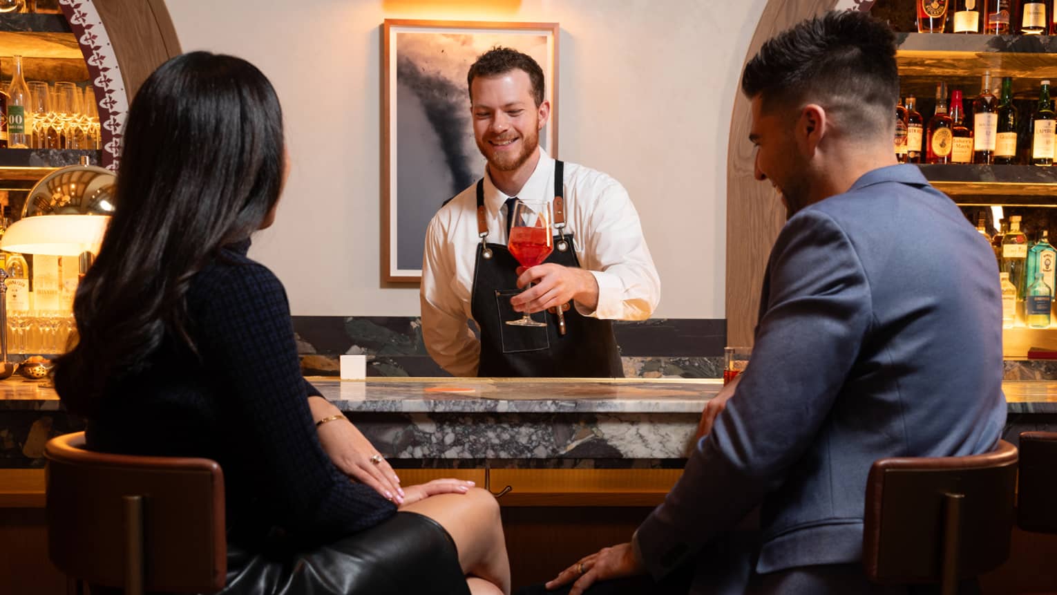 Two guests sitting at a bar while a bartender serves a drink