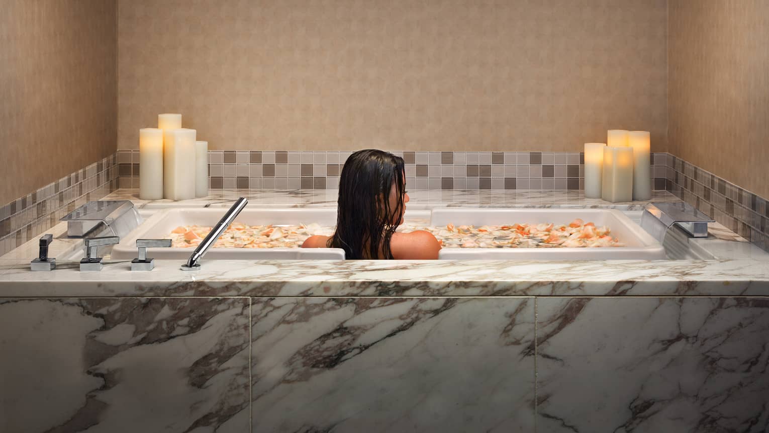 Woman soaks in marble tub filled with flower petals and lined with candles