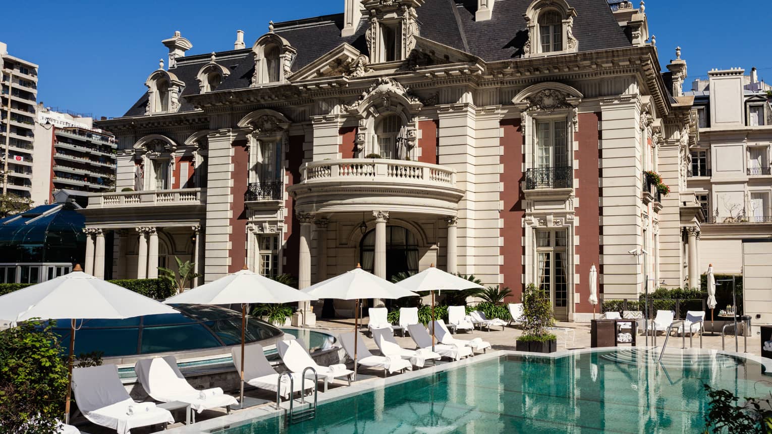 Outdoor pool surrounded by white lounge chairs and umbrellas with a red-and-white 19th century building in teh background