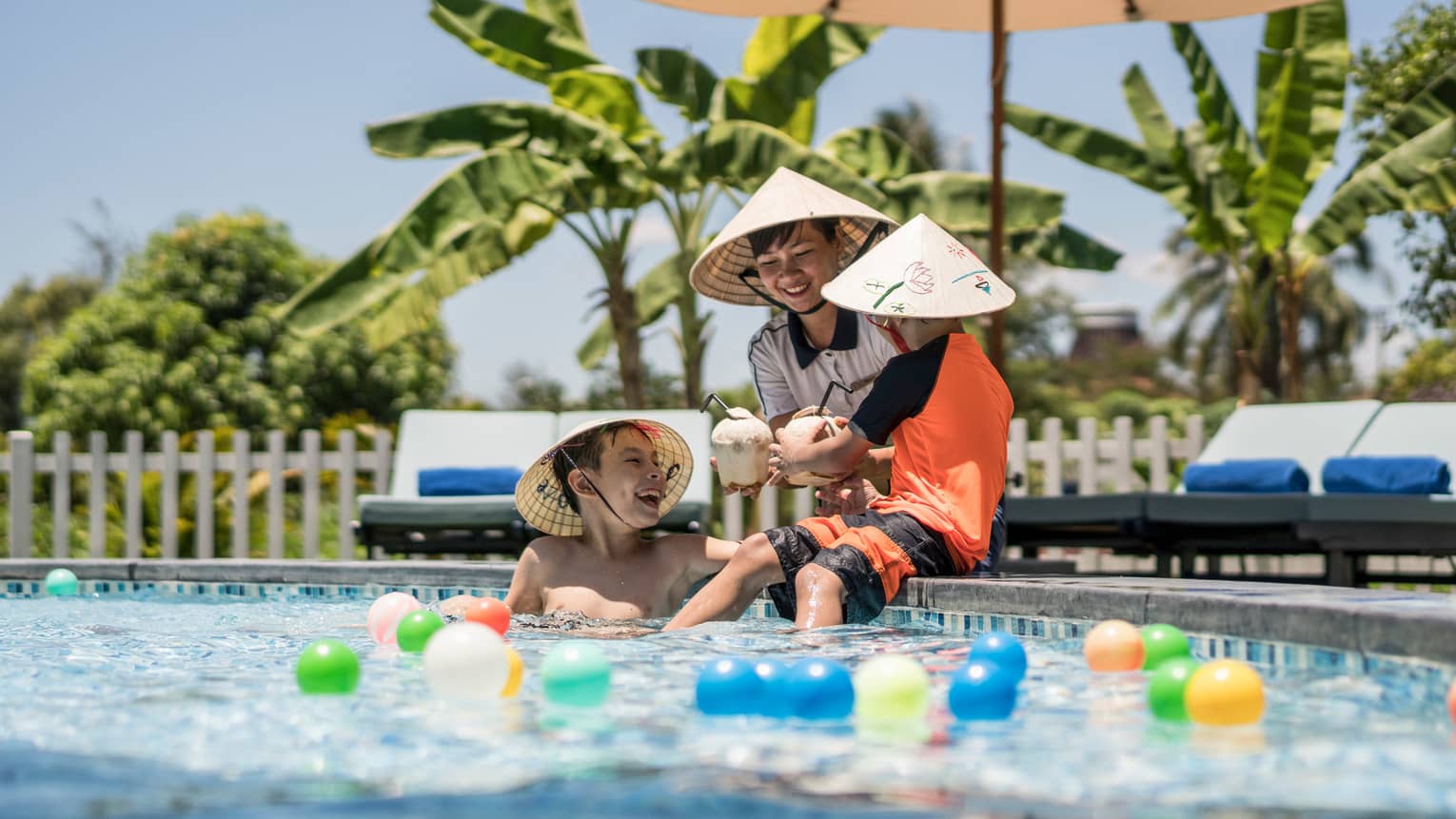 Three laughing children wearing hats, holding coconut drinks play in pool with colourful balls