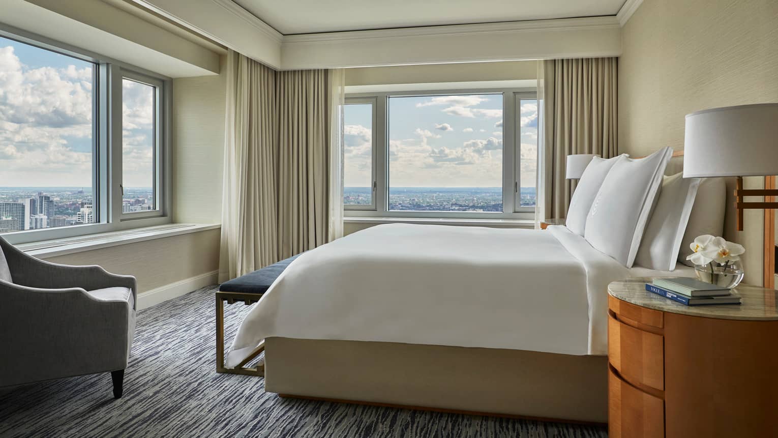 City-View corner suite bed with blue velvet bench, grey armchair, large windows with city views