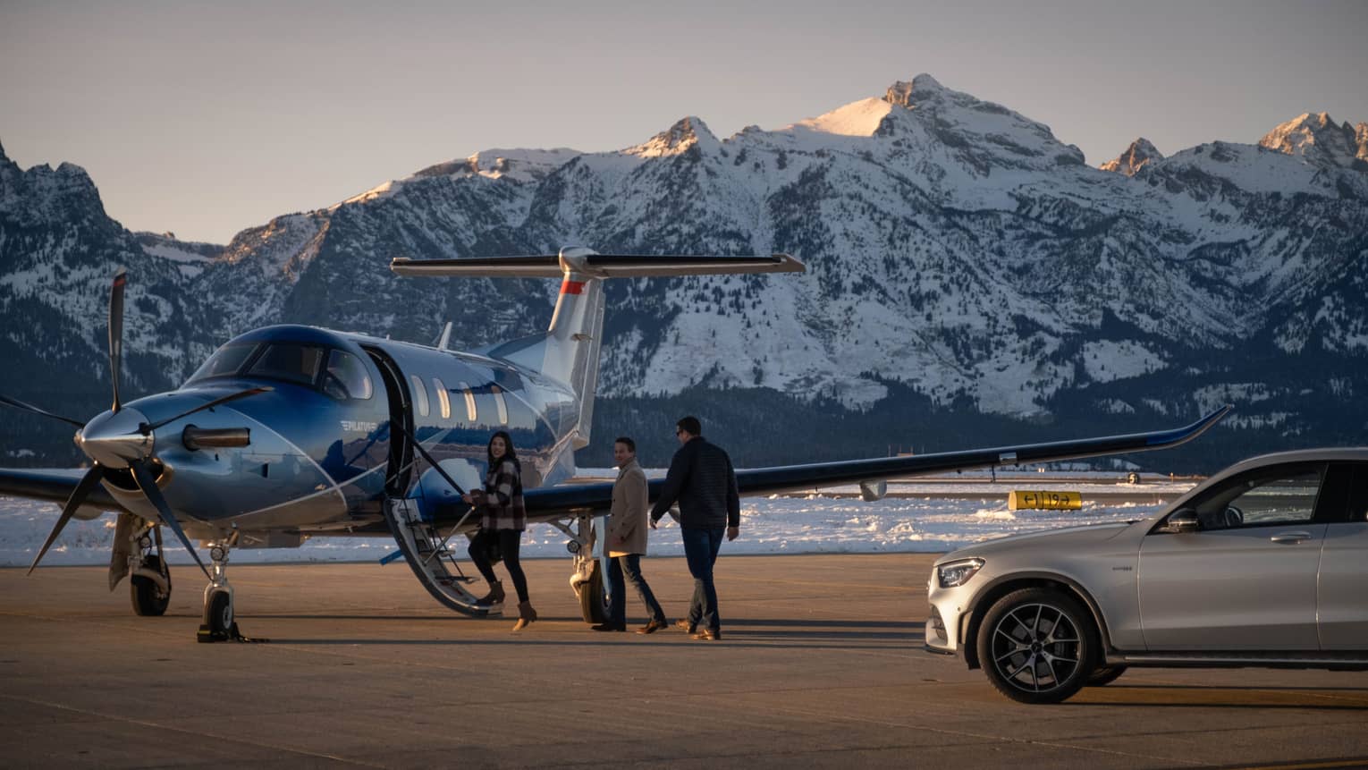 Three people wearing winter coats are heading from a car to a small plane, with snow cover mountains in the distance.