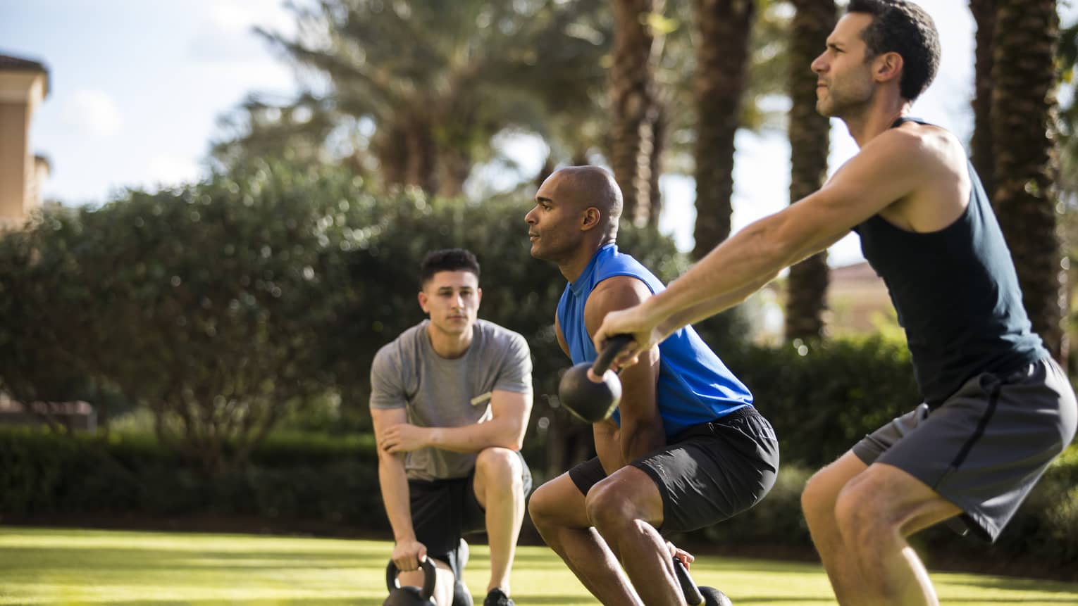 Four Seasons trainer watches as two men squat, lift kettlebell weights on lawn
