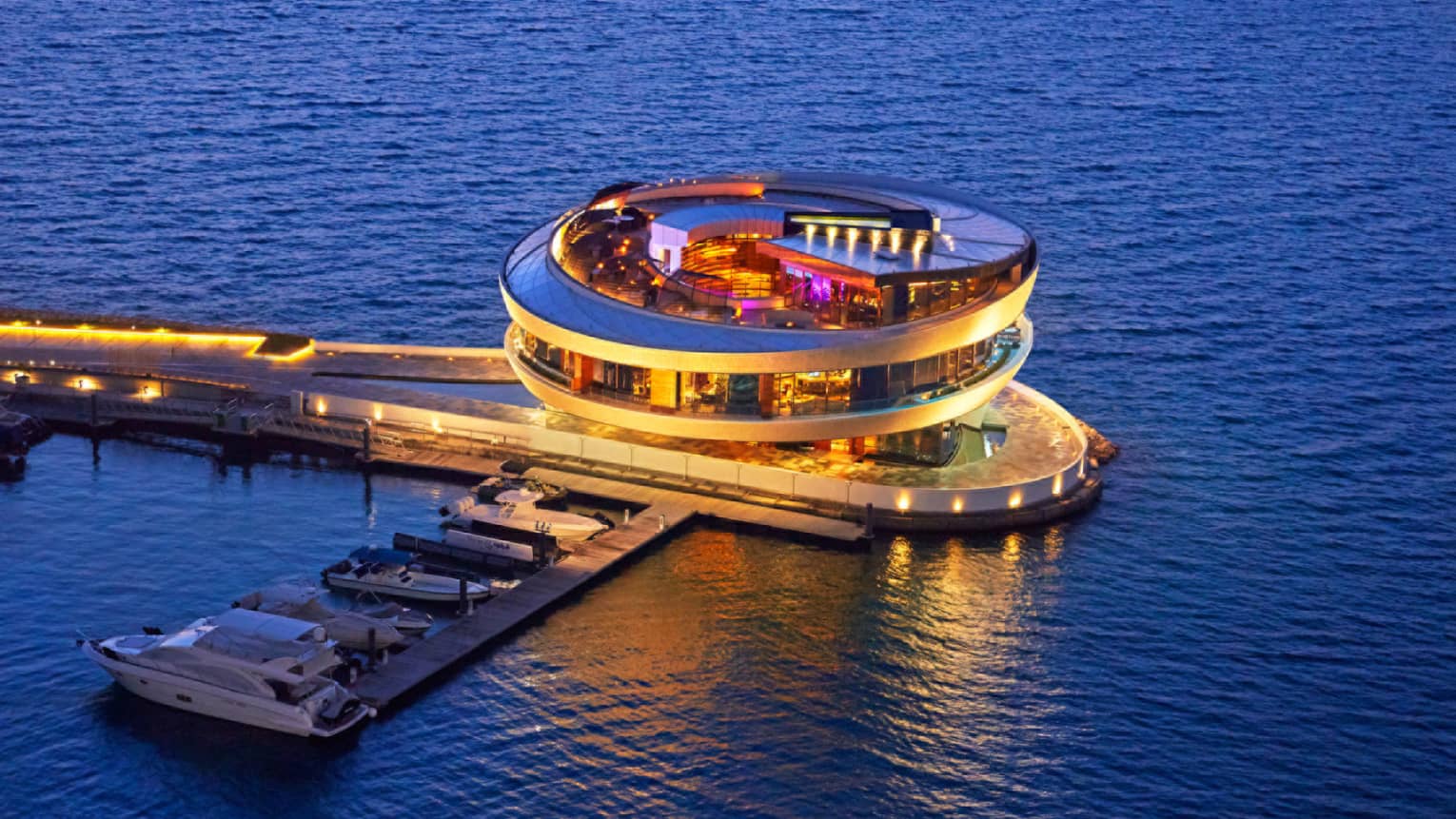 Aerial view of a cylindrical building with vibrant lighting and windows on all sides, on a lit-up pier in deep blue ocean.