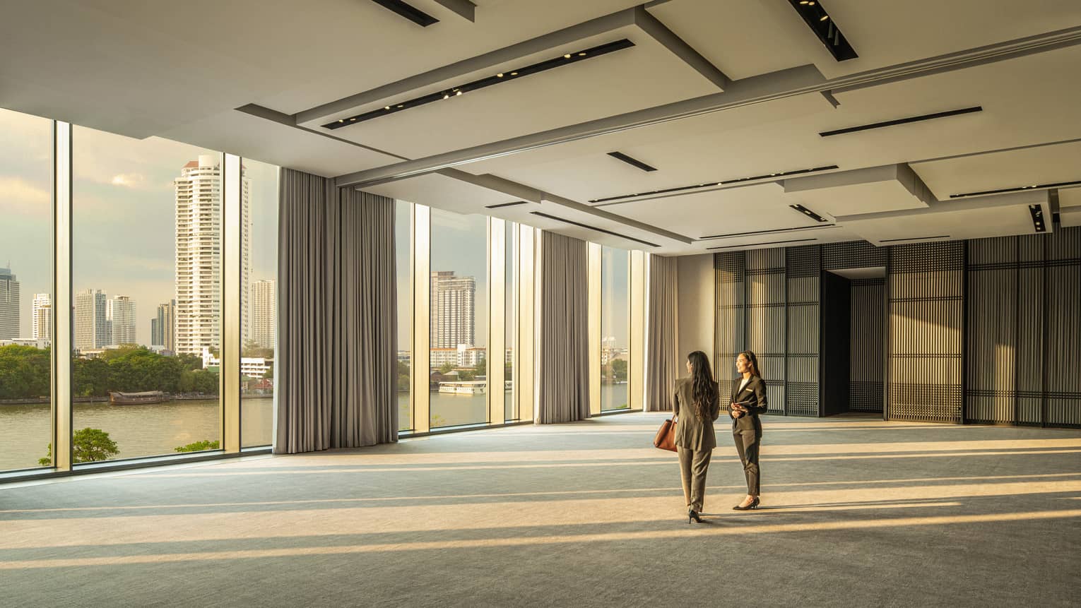 Two woman stand in the empty Conservatory Ballroom gazing out the floor-to-ceiling windows at the Bangkok skyline 