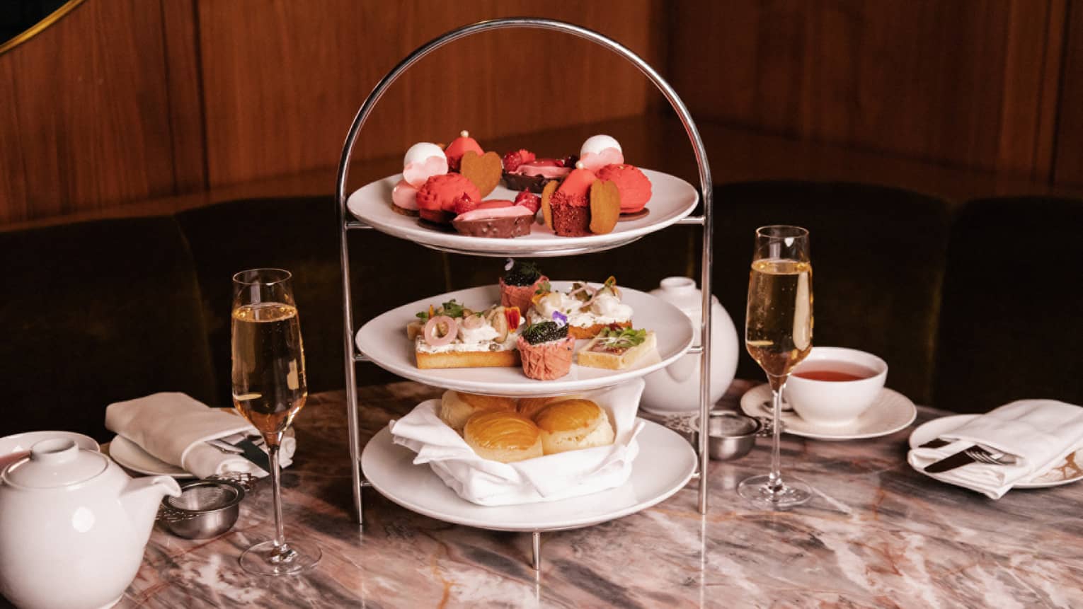 Three-tiered afternoon tea tray filled with pastries and snacks set on a marble table next to two glassed of champage, two cups of tea and a tea pot