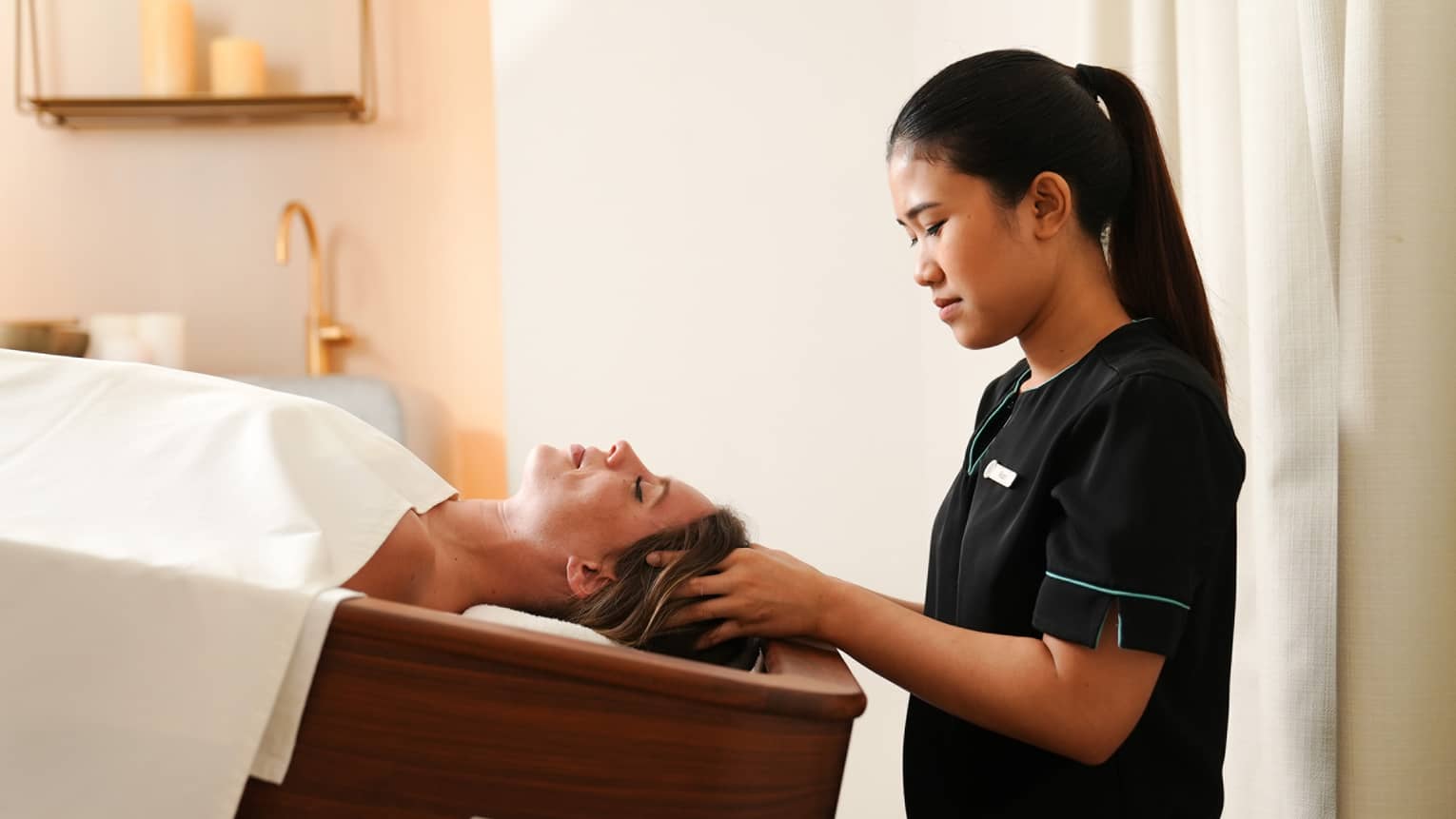 Spa therapist wearing black short-sleeve uniform massages the scalp of a guest laying under a white blanket on an inverted massage table