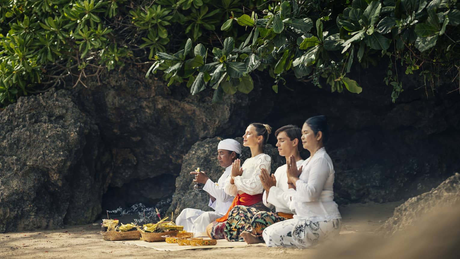 Four adults sit on the ground beside a rock face with their eyes closed and hands held together in front of their chests.