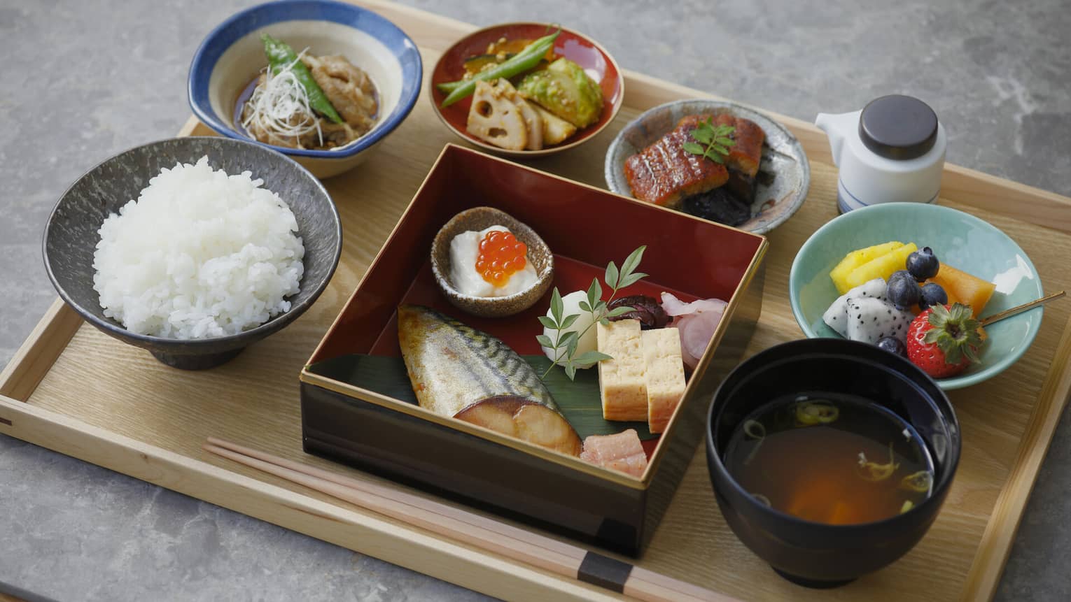 A close up of a Japanese breakfast on a natural wooden tray ? fish, chopsticks, rice, soup and fruit