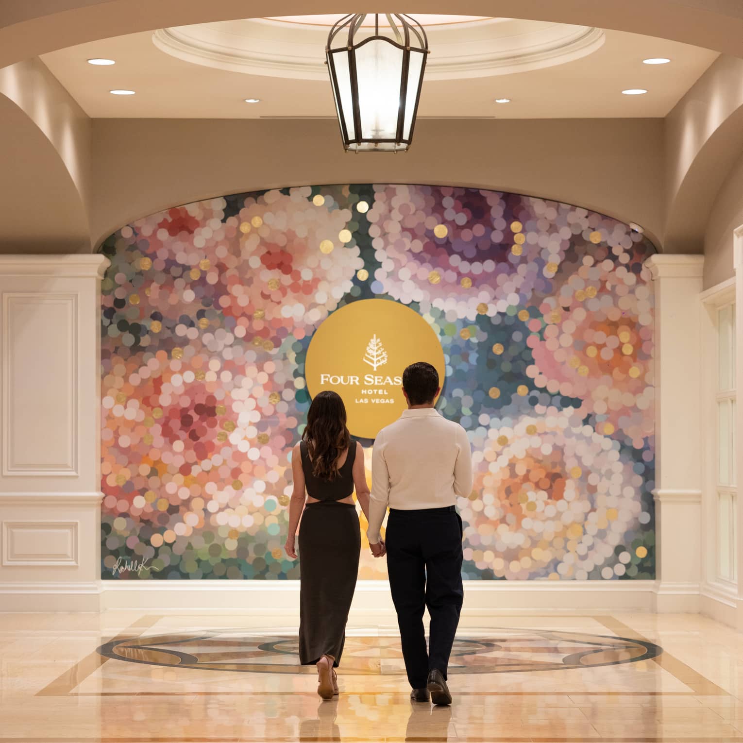 A couple dressed in formal attire hold hands while walking towards a colourful mural in an ornate hotel lobby