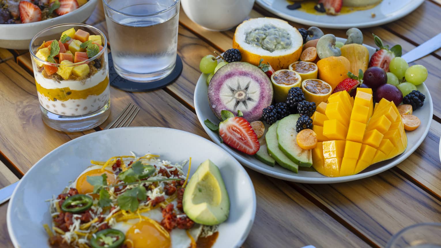 Table filled with plates of colourful food including fresh sliced fruit, sunny-side up eggs, avocado, yogurt bowls and more
