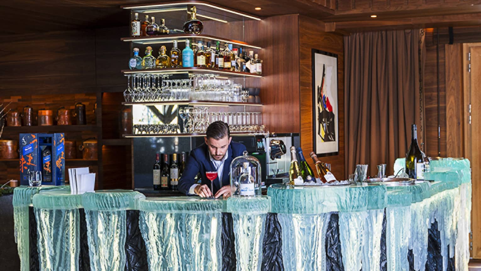 In a wood-panelled room, a bartender in a suit carefully adjusts a cocktail on a glass bar that looks like icicles.