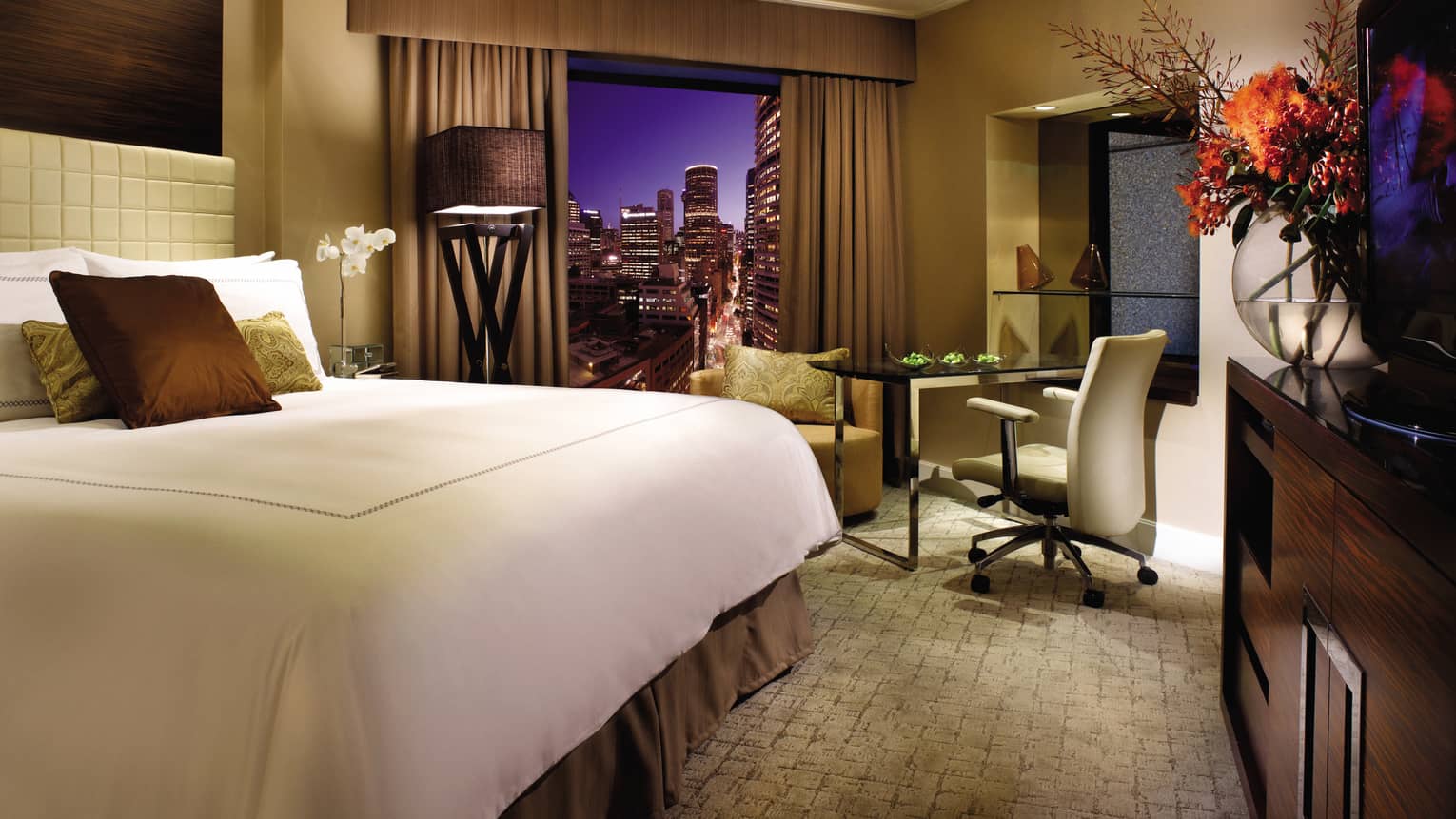 Deluxe City Room at night, bed with brown silk pillows, large glass vase with flowers, window with city view