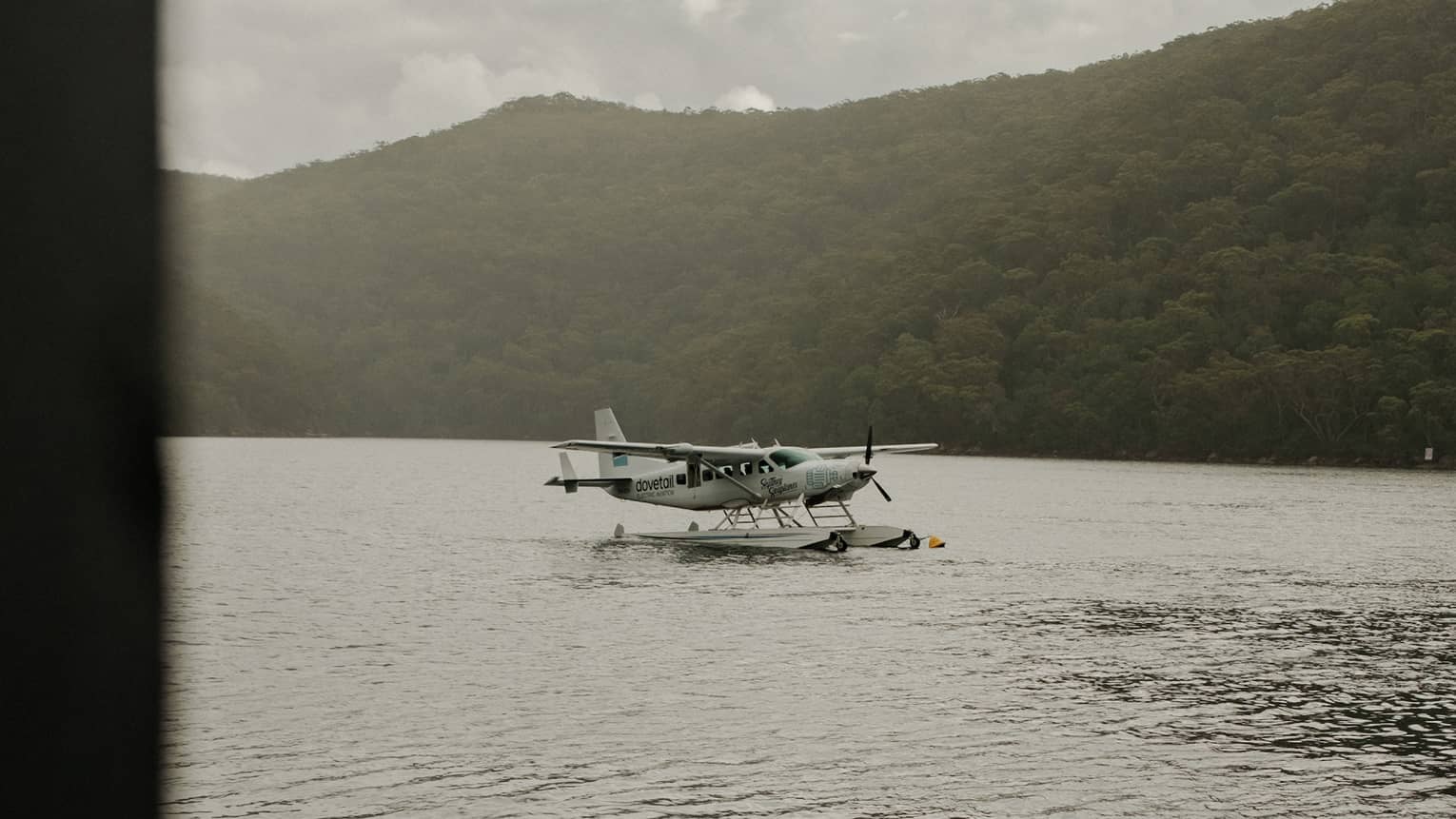 A white seaplane taxiing on water, seen from a sloped onshore gangplank and dock, with lush hills across the water.