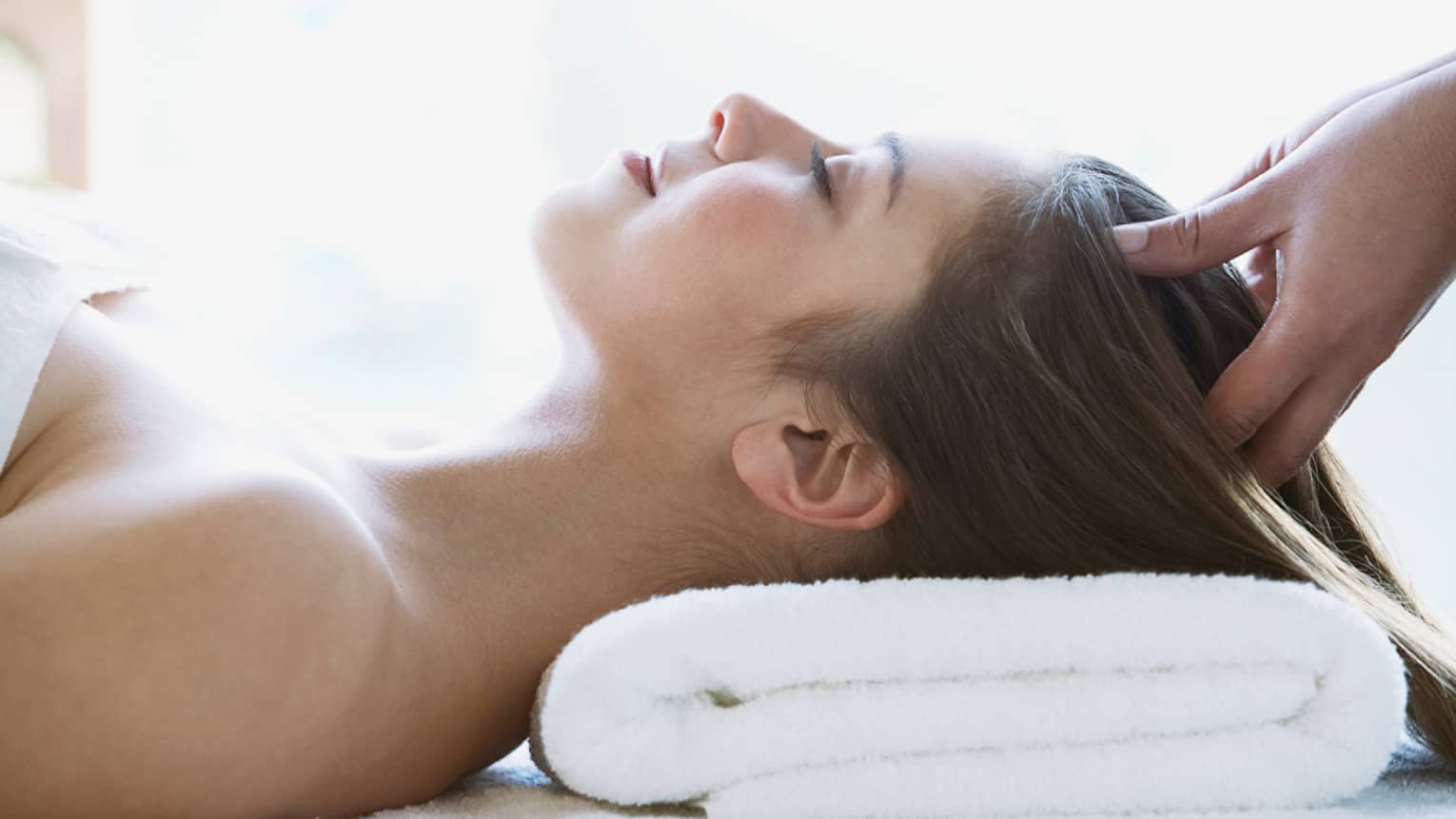 Woman lays on back with head on folded white towel, closes eyes as hands massage her scalp