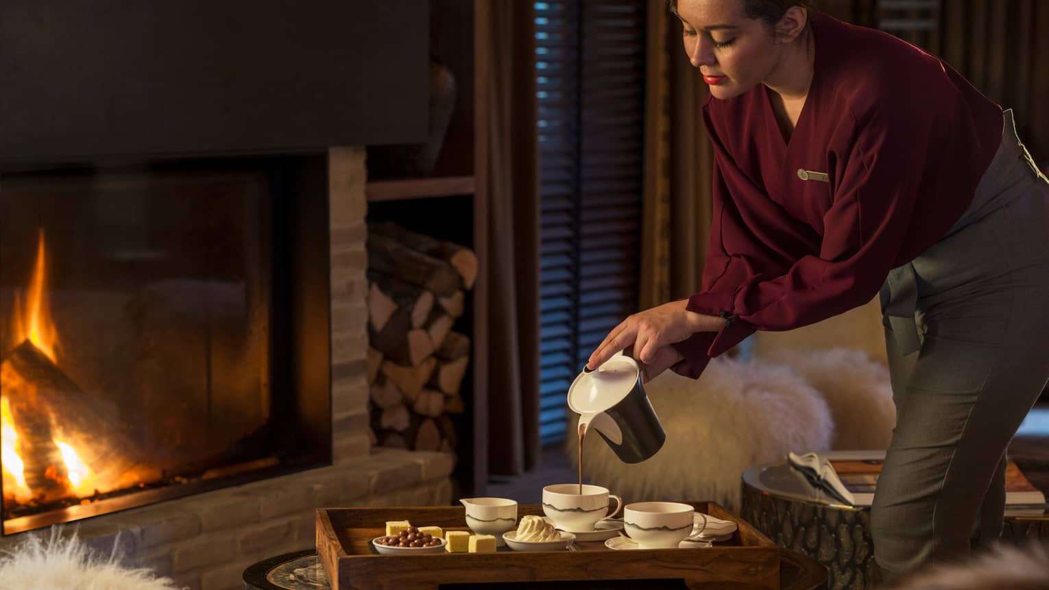 Hotel staff pours hot chocolate into cup on tray with chocolates. marshmallows, cream by fireplace