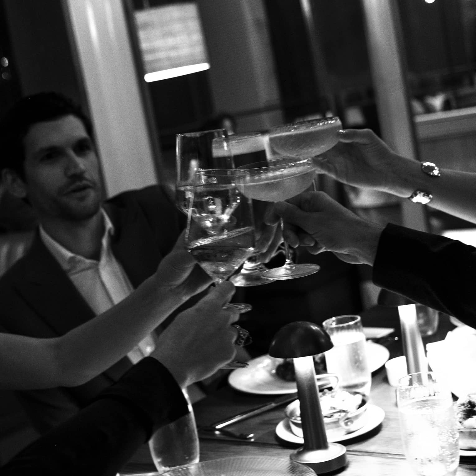Black and white photo of friends raising their wine and cocktail glasses to cheers around a dimly lit restaurant table