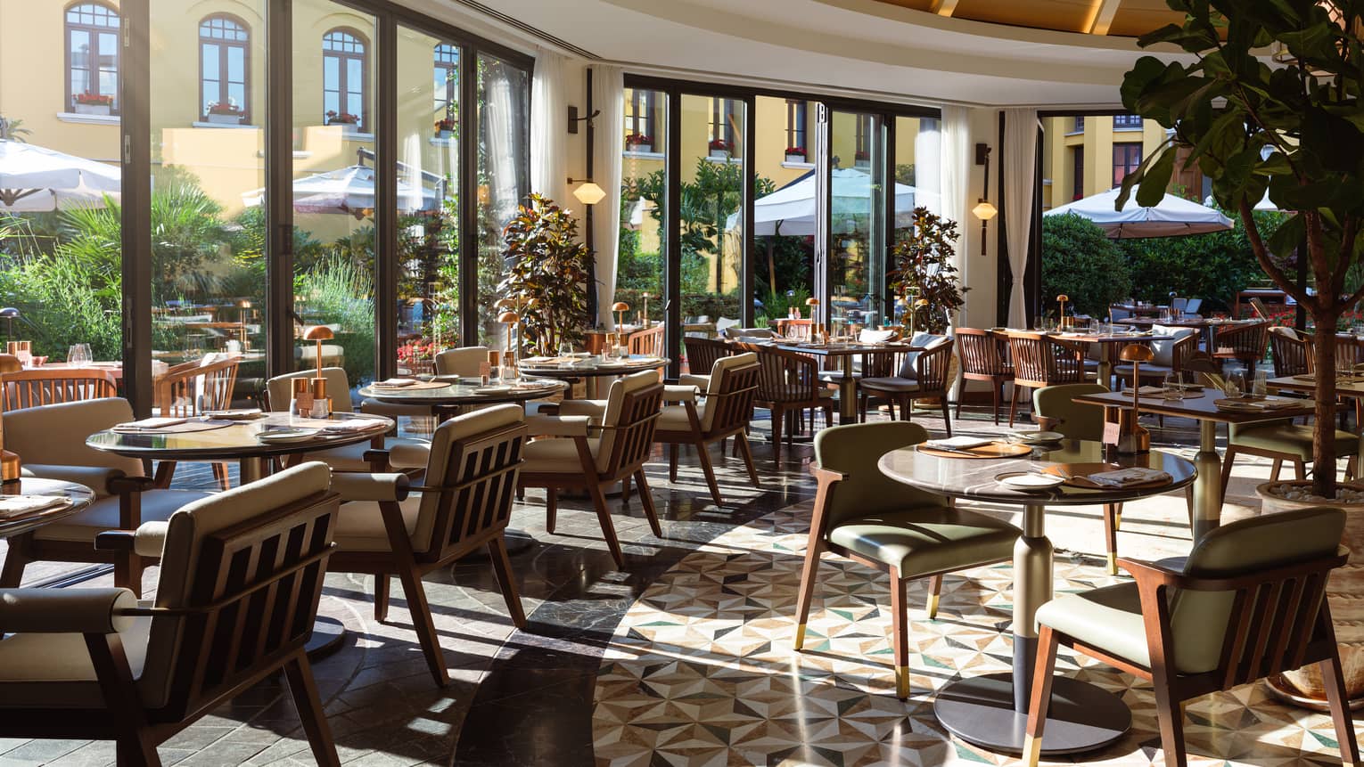 A light-filled indoor dining space with floor-to-ceiling windows along a curved wall, letting in sunny views of the outdoor dining area. Small tables with upholstered seating fill the space.