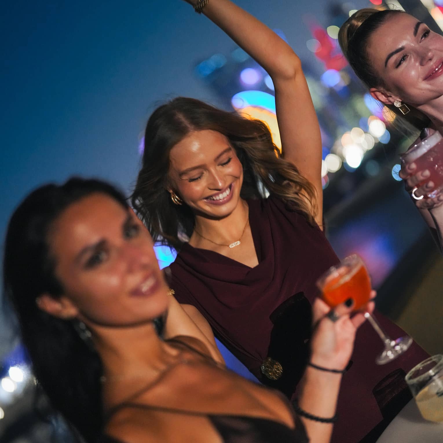 Three guests smile and dance around a cocktail table at a rooftop bar