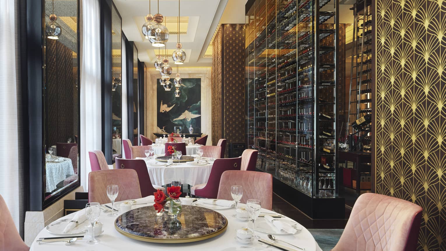 Opulent dining room with gold and pink accents, iron wine rack covering one wall, mirrors opposite, and a painting of cranes.