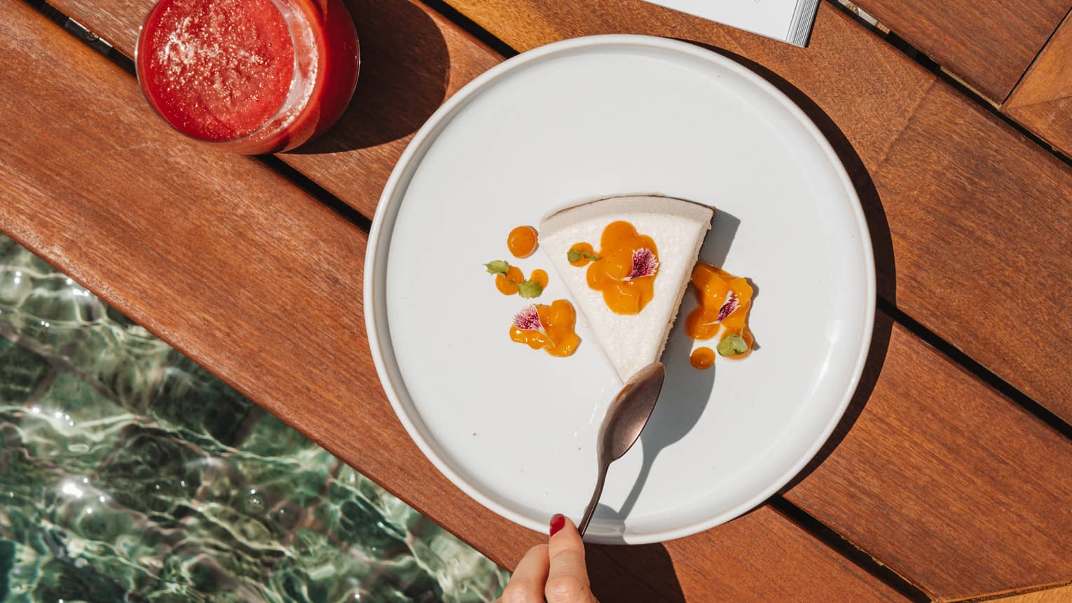 A wood surface with an open book placed on it next to a reddish drink in a tumbler and a white plate holding a white and orange food item. A person reaches for a bite of the food with a spoon.