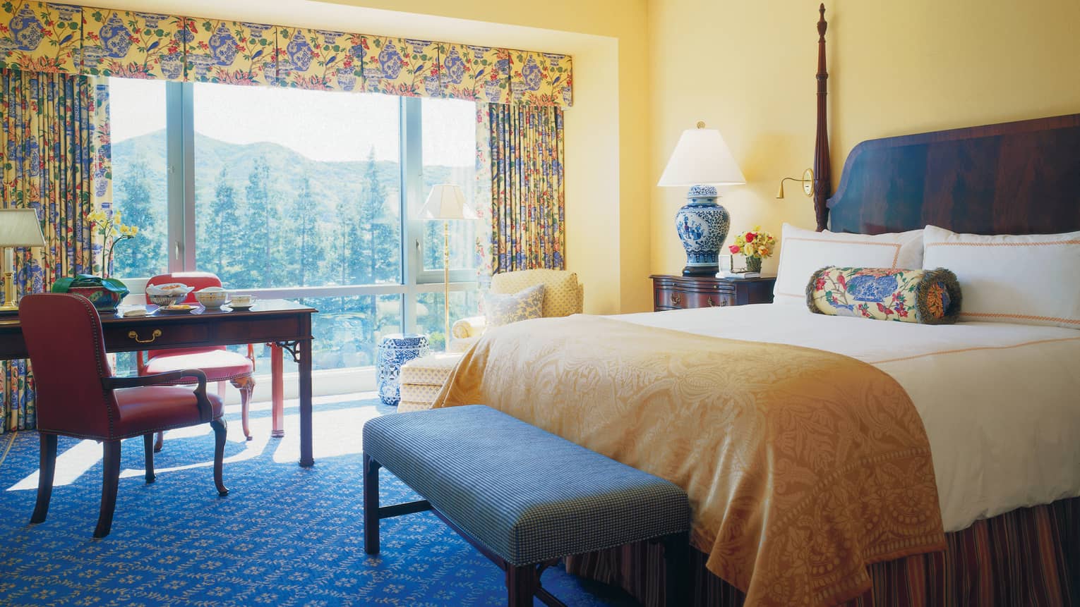 Deluxe Room hotel bed with gold blanket, bench, desk, windows with bright curtains