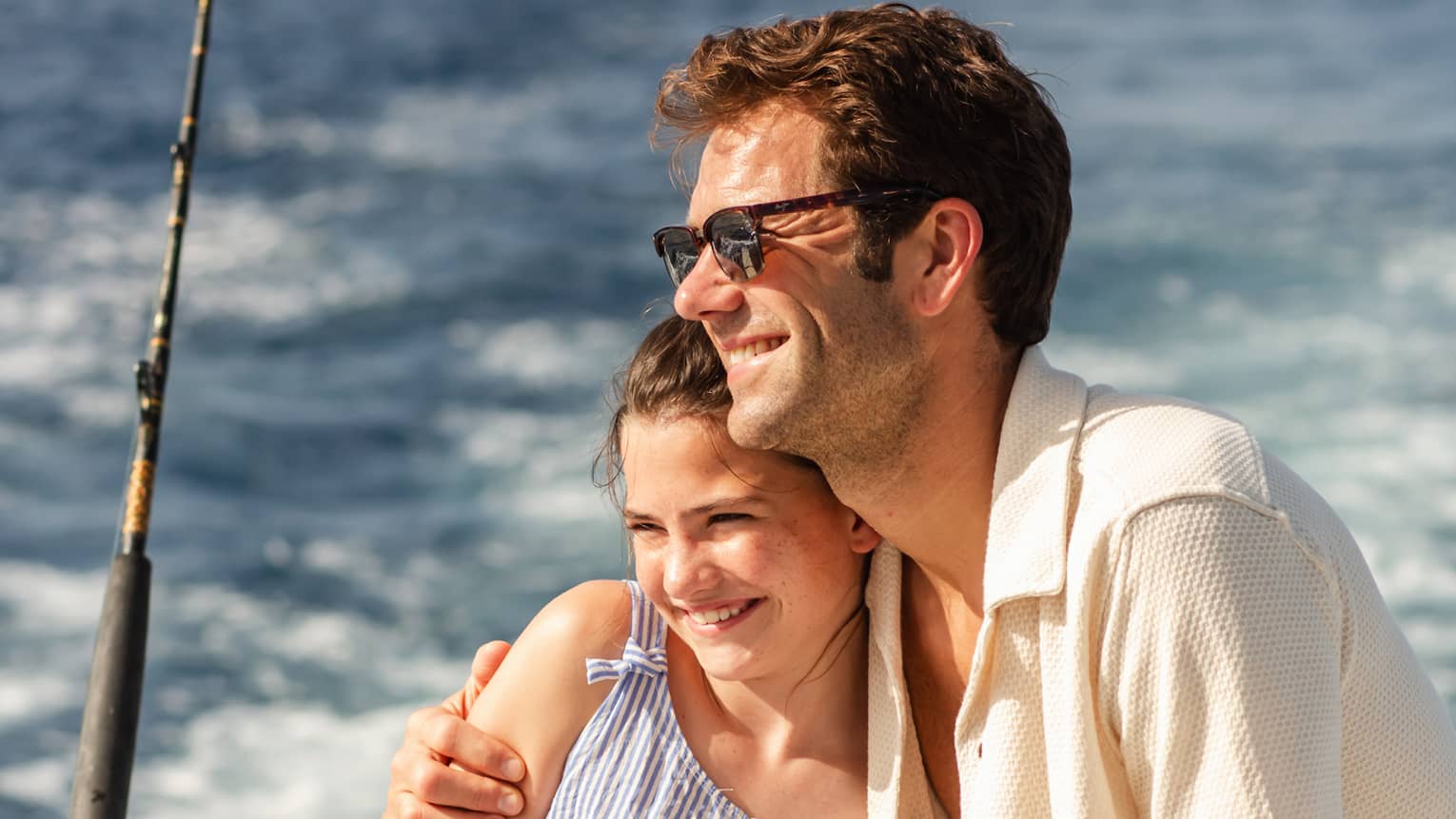 Onboard a boat, two smiling guests hug in the summer sunshine, an upright fishing rod attached to the boat's edge nearby.