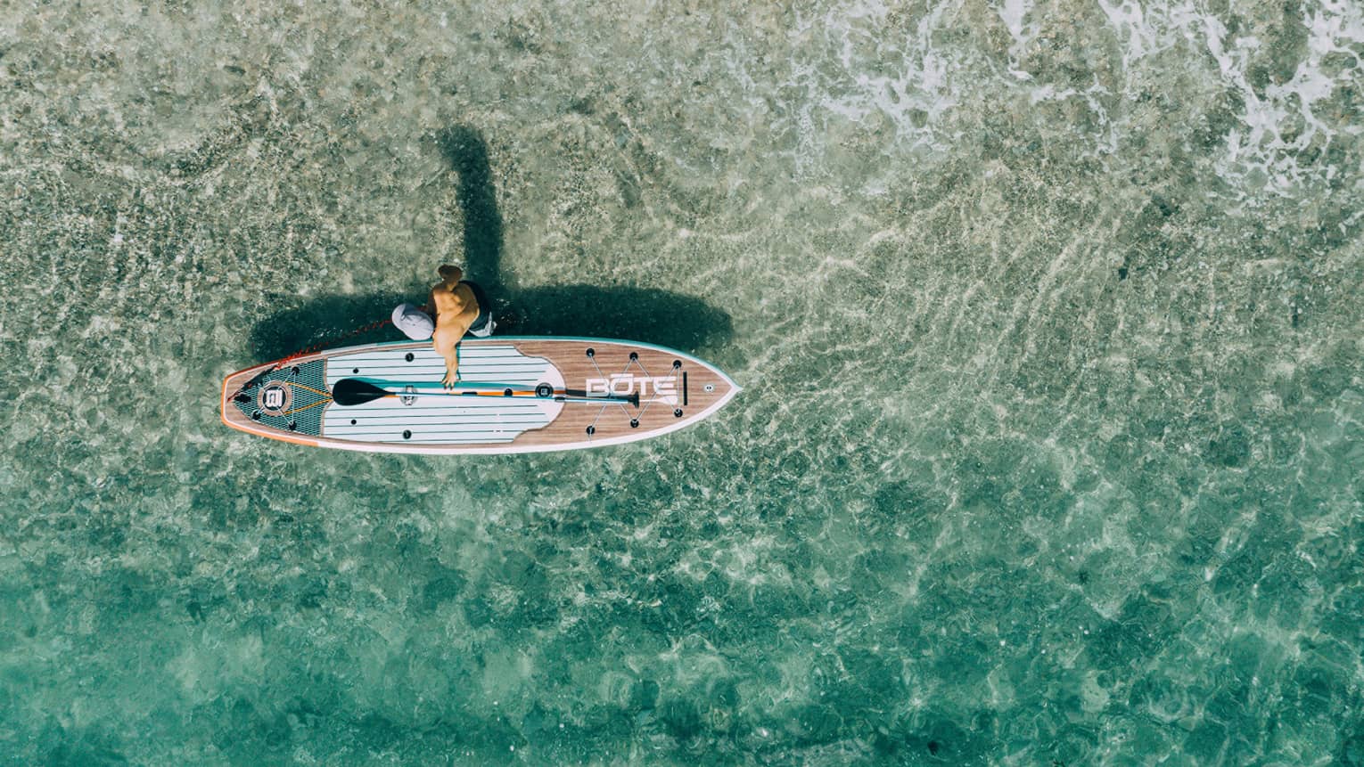 Bird’s-eye view of a paddleboarder walking beside his board in shimmering, crystal-clear water.
