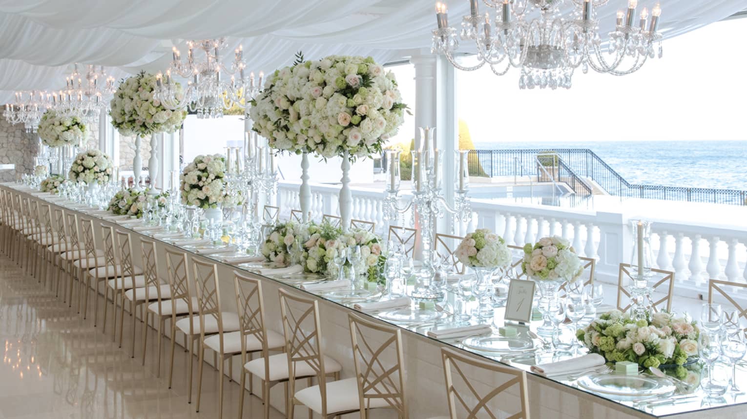 Long banquet table lined with gold chairs, formally set table, white topiaries, crystal chandeliers, sea view