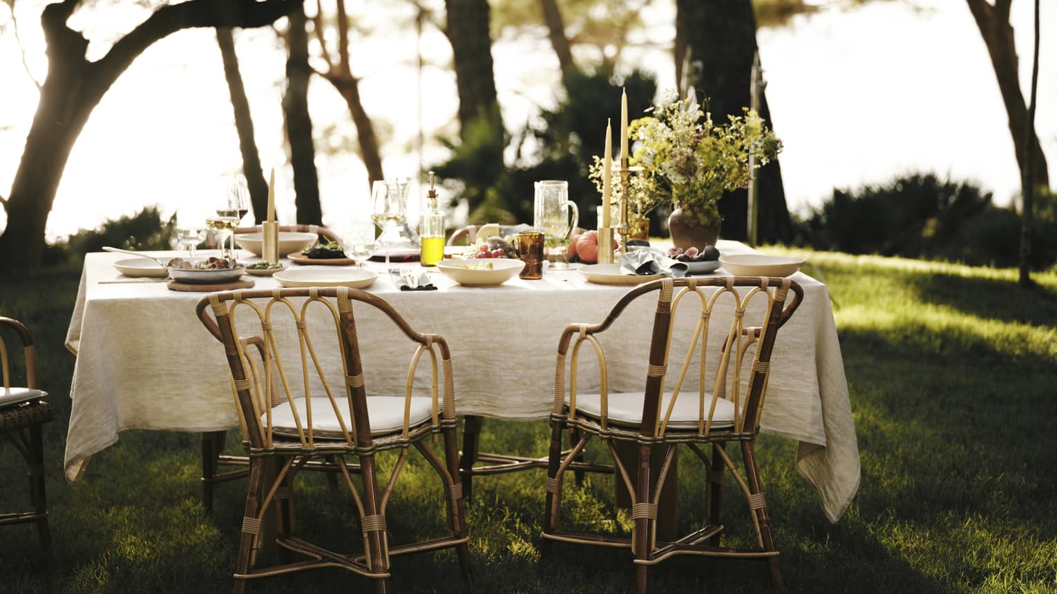 A linen tablecloth adorns an outdoor table set for five with wine, fresh seafood and fruit in an ocean-view forest setting.