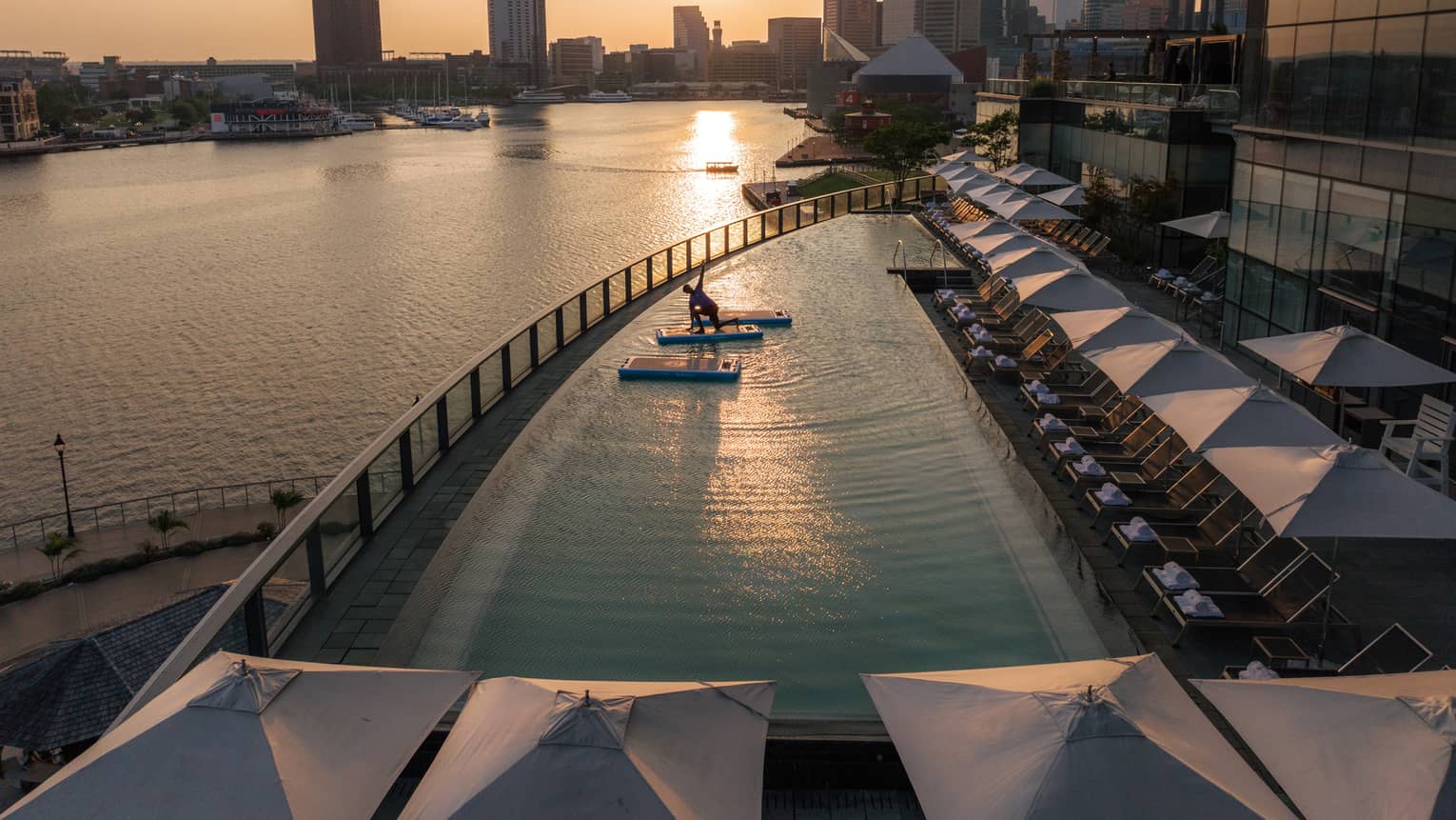 A guest practises yoga on a mat floating in a pool overlooking a river. In the background, the sun sets behind skyscrapers.