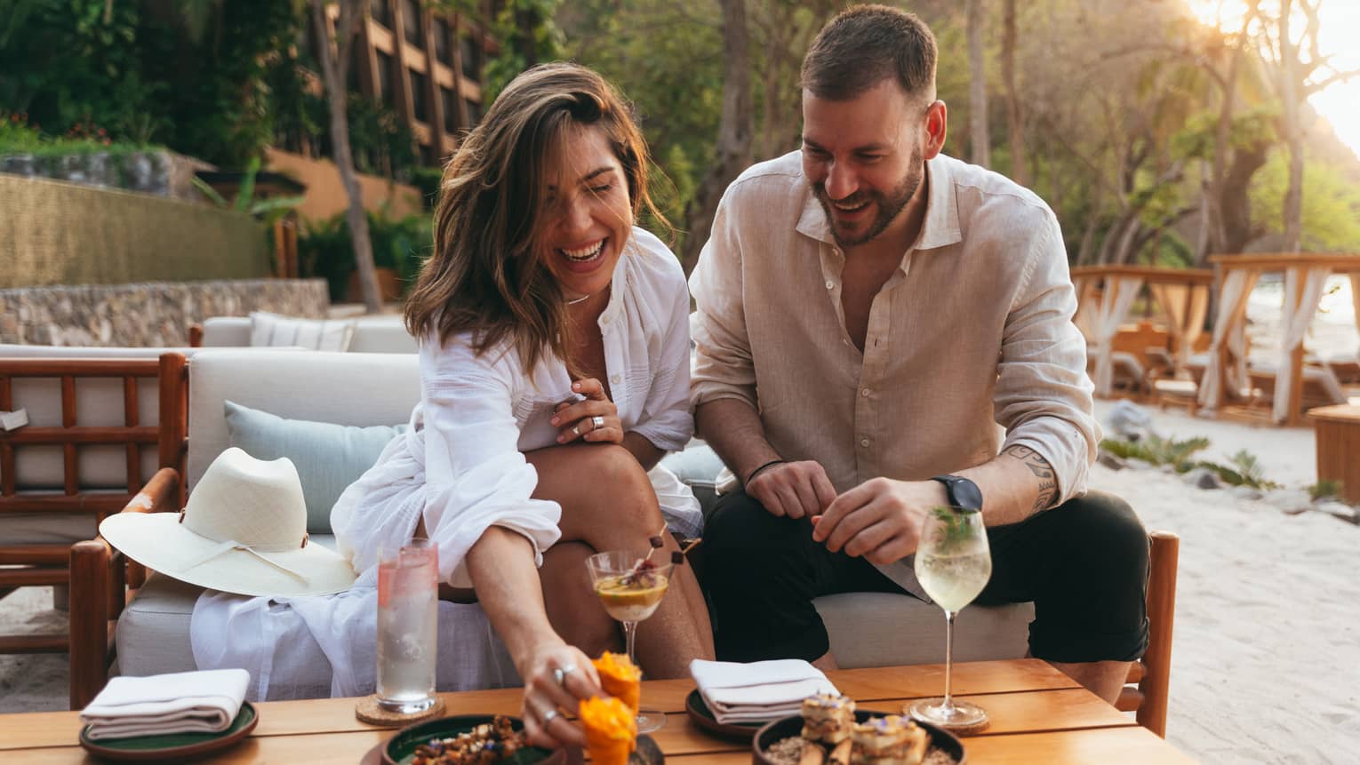 A couple sits a beachfront table smiling and enjoying food and drink