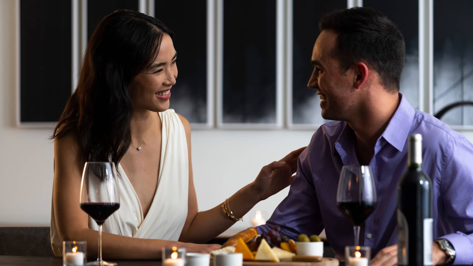 A couple exchanges smiles as they enjoy a cheese platter and a bottle of wine.