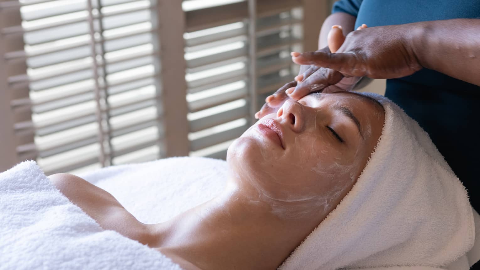 Woman gets facial. A white towel protects her hair as white cream is massaged into her face.