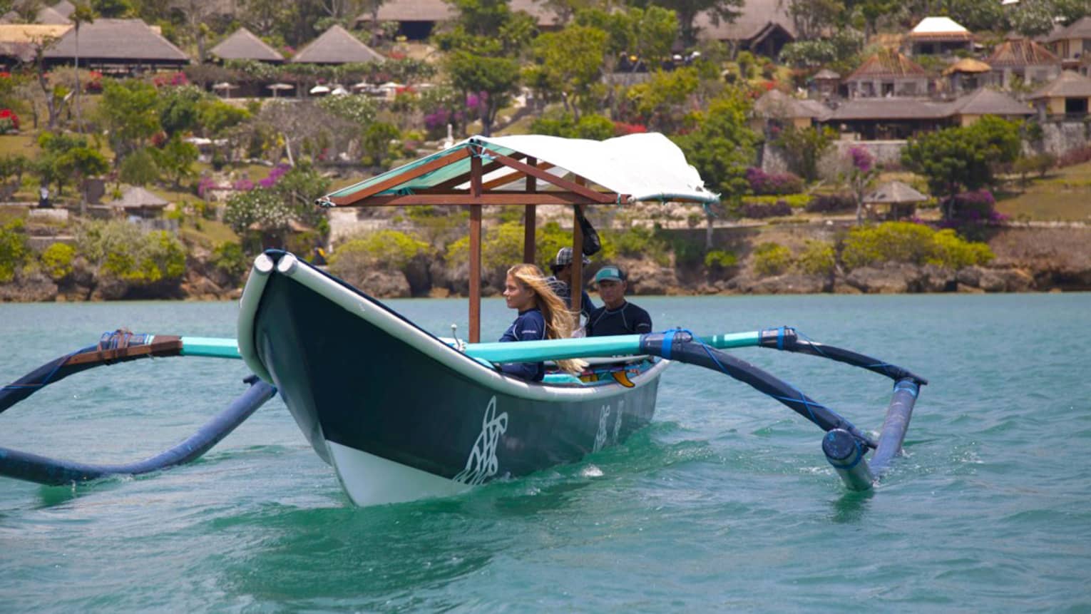 A guide sits at the stern of a covered outrigger canoe as two guests gaze at the water; behind, trees and huts dot the shore.