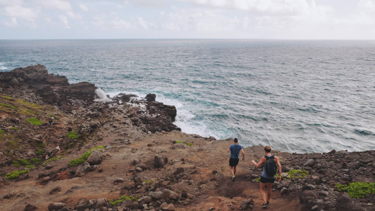 Two people hike down dirt-covered path, rocks towards ocean