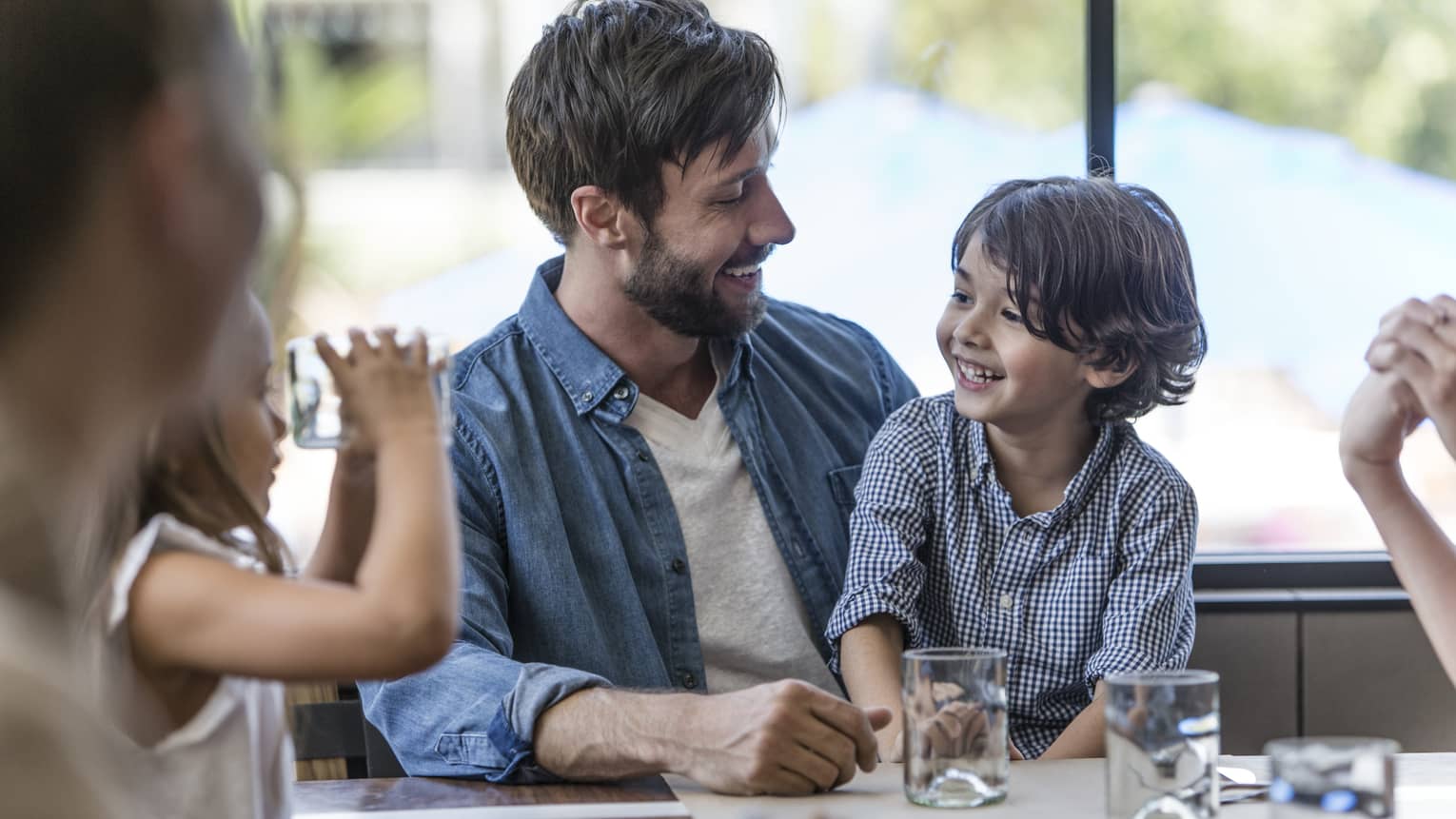 A child sits in his father’s lap and they smile at one another as another child looks at them through the bottom of a glass.