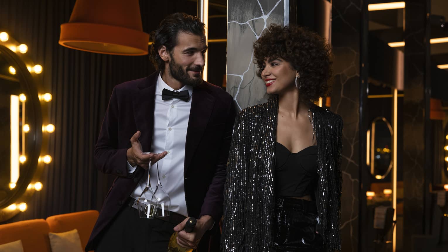 A man in a tuxedo holds a bottle of champagne and two empty champagne flutes as he talks to a woman wearing a black dress and a shimmering black jacket