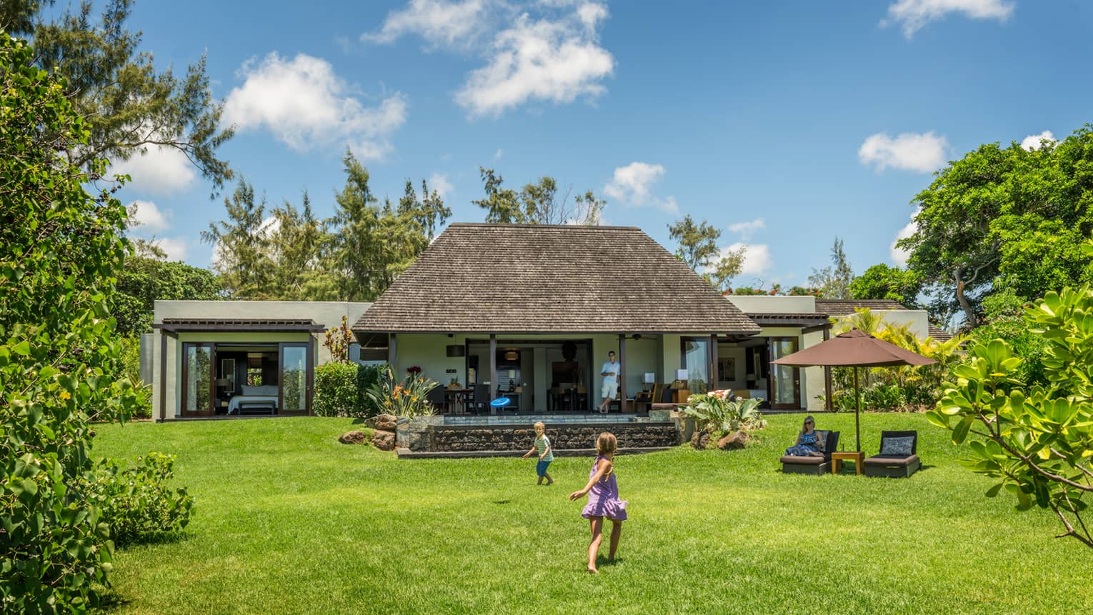 Young children play on green lawn in front of bungalow as dad watches from covered patio