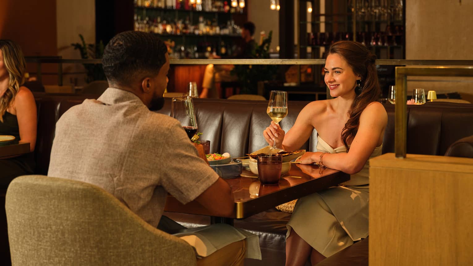 Two people drink beverages from stemmed glasses, sitting across from each other at a dining table in a serene and cozy indoor dining space.