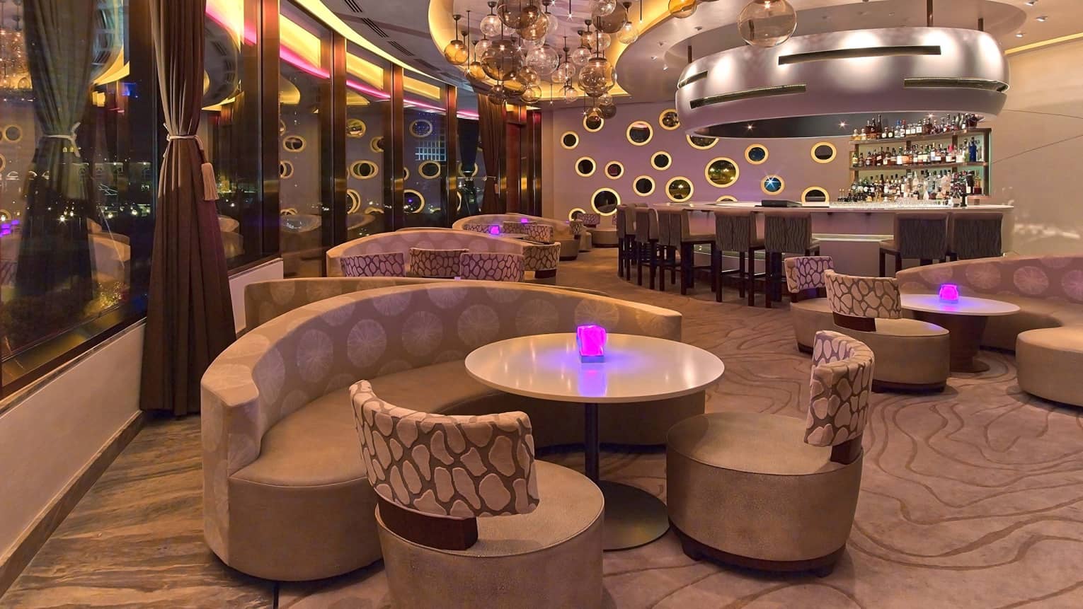 Nighttime interior of Nobu Doha featuring curved seating, cocktail tables, round glass lighting fixtures hanging from the ceiling and pink, purple and yellow lighting accents