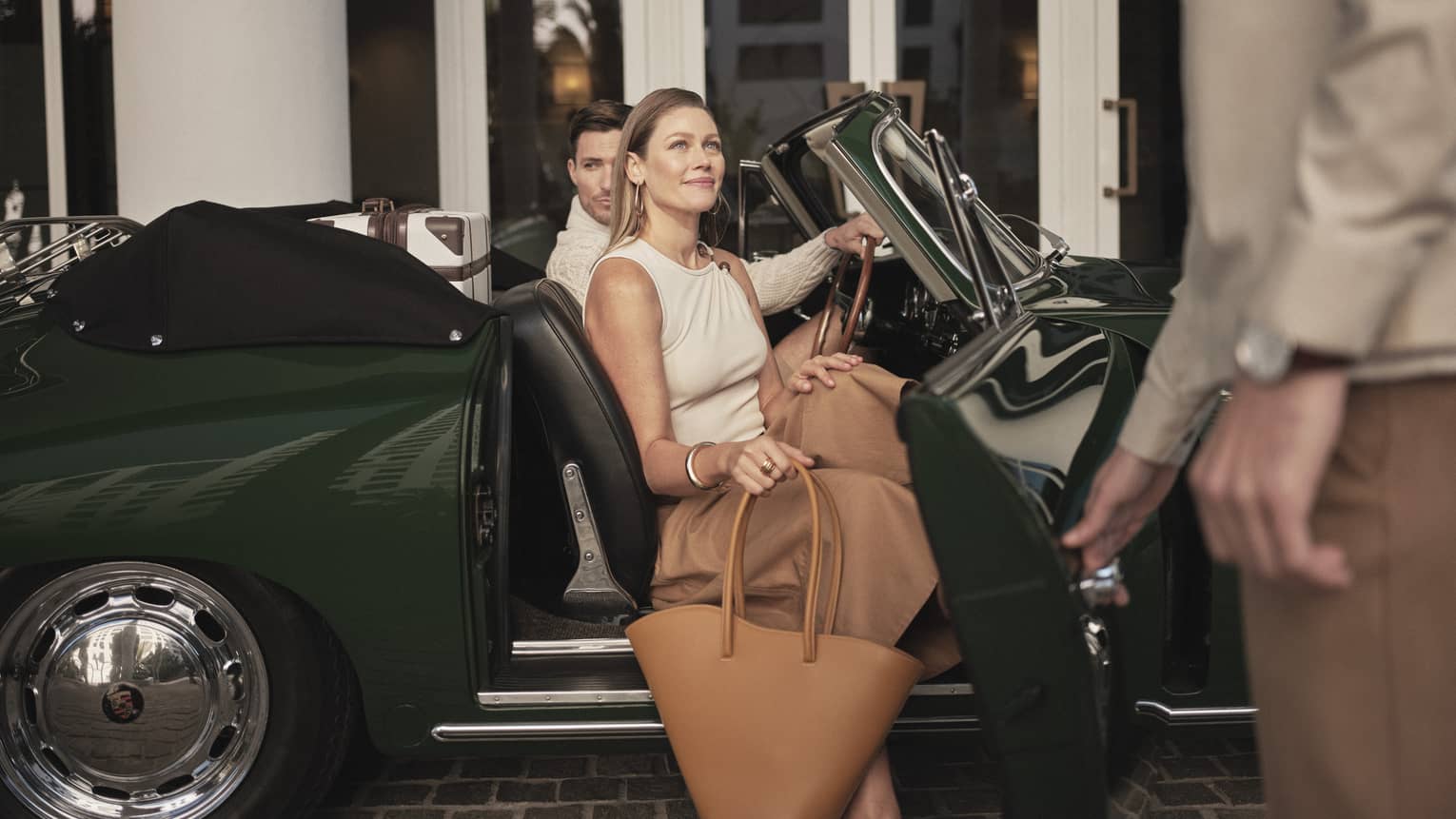 A man and woman getting out of a dark green car holding bags, two hotel team members are assisting them.