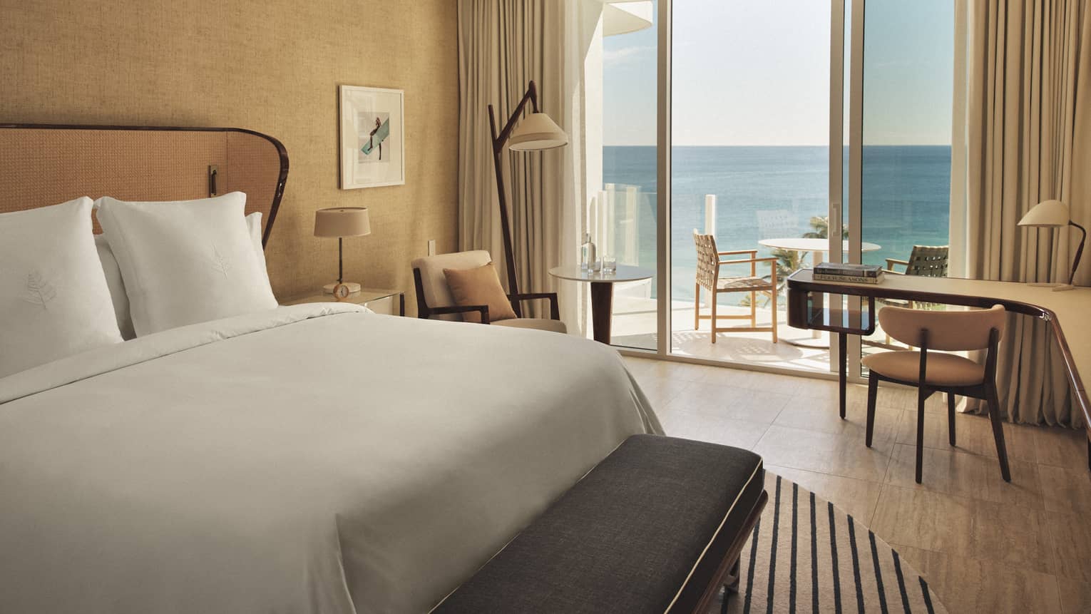 A hotel room with sliding glass door, bed with white sheets, long curved desk, two lamps, two chairs and outdoor seating.